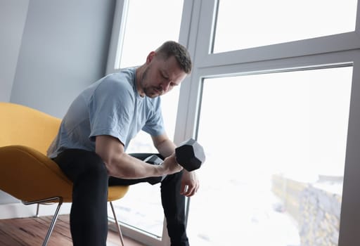 Young man sitting on chair and lifting dumbbell. Home sports training during self-isolation concept