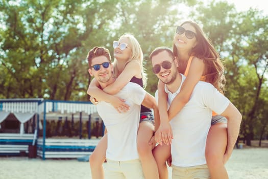 Group of friends walking along the beach, with men giving piggyback ride to girlfriends. Happy young friends enjoying a day at beach.