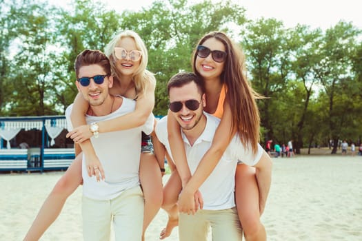 Group of friends walking along the beach, with men giving piggyback ride to girlfriends. Happy young friends enjoying a day at beach.