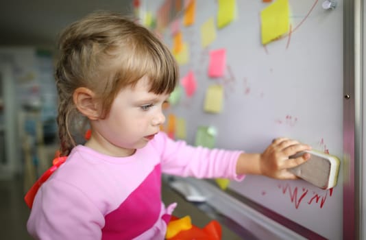 Portrait of smart small girl standing in modern classroom and holding special erasing marker utility to start lovely drawing again. Creative childhood concept