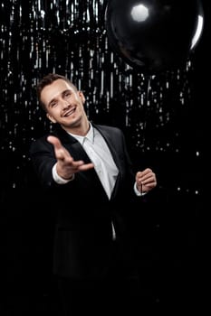 Happy man in black jacket with black air balloons points at camera choosing you on glitter background.
