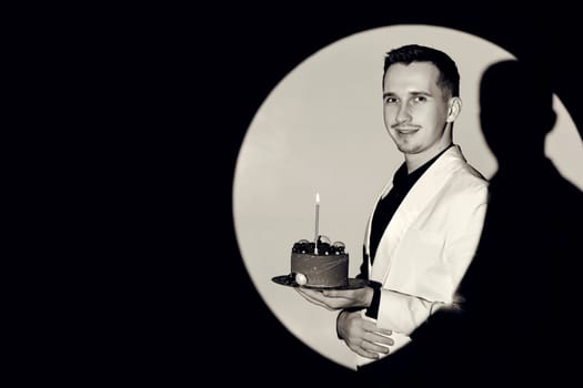 elegant caucasian man in white suit tuxedo holding cake with candle in the circle of light, copy space. Celebration