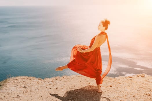 Side view a Young beautiful sensual woman in a red long dress posing on a rock high above the sea during sunrise. Girl on the nature on blue sky background. Fashion photo.