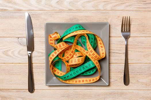 Top view of colorful measuring tapes on plate in the form of spaghetti with knife and fork on wooden background. Weight loss and diet concept.