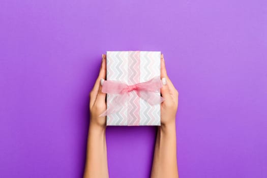 Flat lay of woman hands holding gift wrapped and decorated with bow on purple background with copy space. Christmas and holiday concept.