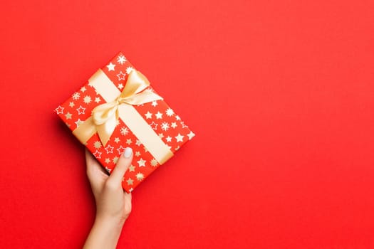 Female's hands holding striped gift box with colored ribbon on red background. Christmas concept or other holiday handmade present box, concept top view with copy space.