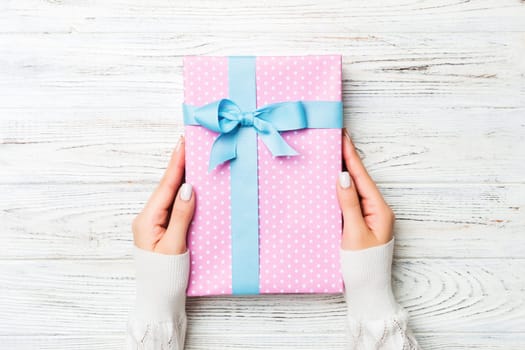 Top view woman hands holding present box with colored bow on white rustic wooden background with copy space.