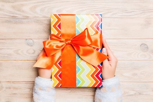 Woman hands give wrapped christmas or other holiday handmade present in colored paper . Present box, decoration of gift on yellow rustic wooden table, top view with copy space.