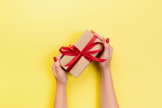 Woman hands give wrapped valentine or other holiday handmade present in paper with red ribbon. Present box, decoration of gift on yellow table, top view with copy space.