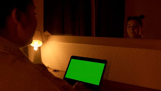 Woman looking at laptop with green screen during night time in front of mirror. Media. Freelancer watching monitor display with chroma key screen, working overtime concept