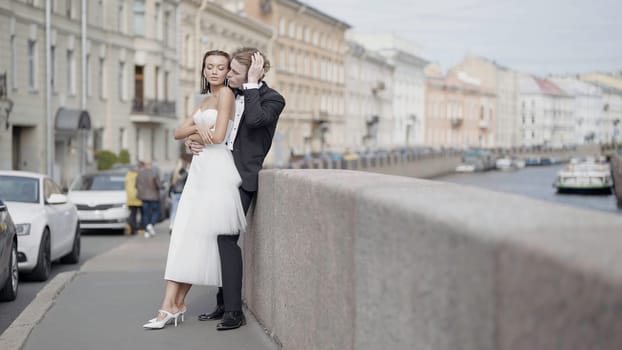 Beautiful newlyweds. Action. A couple of lovers, the bride in a tight white dress and the groom in a suit with long hair pose on the street next to the embankment and beautiful historical buildings in summer