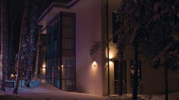 Facade of the big house in the late winter evening illuminated by colorful flashing light of an emergency car. Outdoors view of the cottage in snowy winter at night.