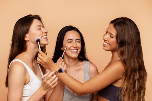 Smiling millennial international women in underwear apply nude makeup with brushes to friend, enjoy beauty care, isolated on beige background, studio. Cosmetics, fashion ad and offer