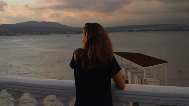 Woman on embankment at sunset. Media. Young woman stands at railing on background seascape at sunset after rain. Beautiful landscape opens up to attractive woman on embankment.