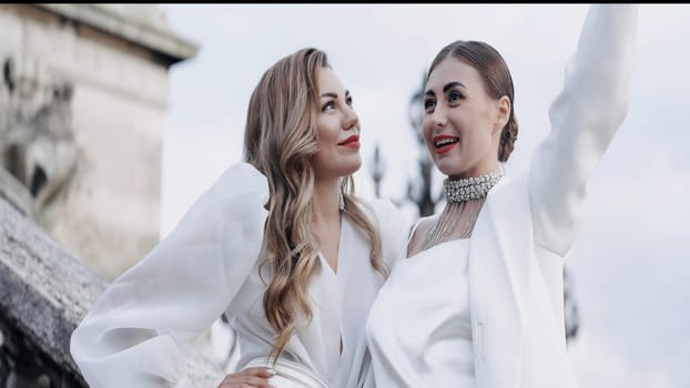 Two beautiful women blondes with red lips posing outdoors. Action. Adult ladies in white clothes holding a hat made of feathers, concept of fashion
