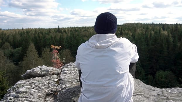 Rear view of a mountain hiker looking into autumn landscape of the forest and cloudy sky. Man sitting on the top of the high rock looking at trees peaks