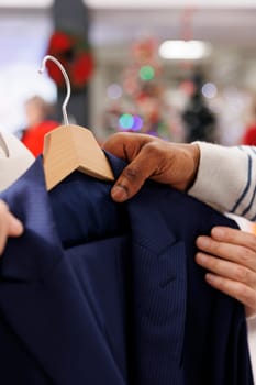 Store worker showing jacket to client in fashion boutique, presenting new merchandise on racks. African american employee describing material on clothes, christmas shopping. Close up.