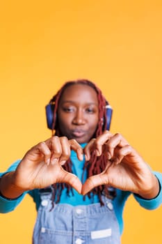 Person doing heart shape sign in studio, listening to music on headphones. Female model showing romantic symbol and love gesture on orange background, advertising romance and flirt.