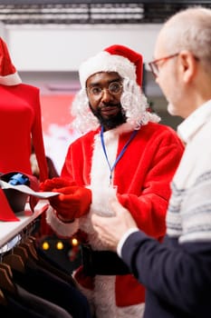 Worker presenting ties to senior man, recommending accessories for christmas eve festivity formal attire. Assistant dressed as santa claus talking to customer about fabric quality and outfits.