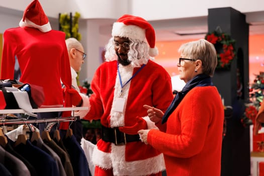 Man in santa costume showing items on racks to client, senior woman asking employee about sizes available in store. Jolly person in red suit helping customer to find xmas gift.
