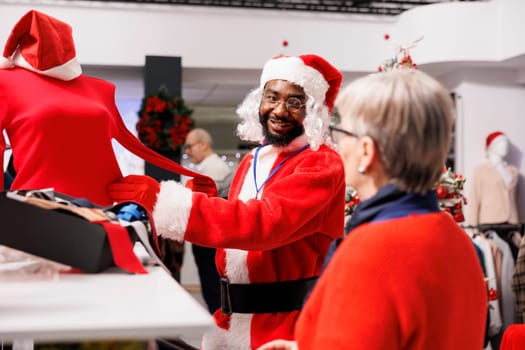 Store employee guiding client in shop, wearing festive santa claus suit while helping customers with christmas present ideas and suggestions. Young man recommending blouse to senior woman.