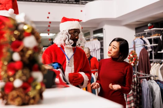 Store worker in santa suit helps client and presenting new clothing collection at shopping mall, talking to asian woman about christmas present ideas. Festive person in costume greeting buyer.