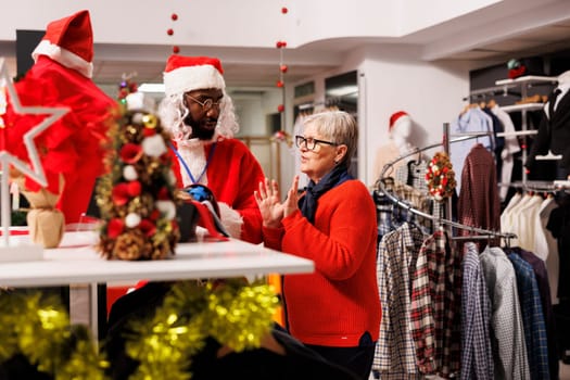 Employee in suit presenting clothes to woman, searching for fashion items as presents. Man portraying santa claus greeting customers and giving assistance during holiday sales.