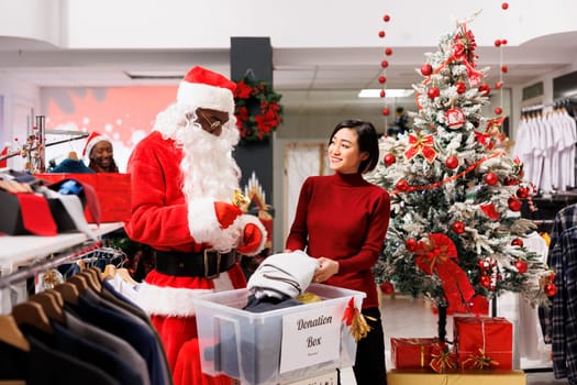 Asian woman helping with charity donations in clothing store, working with santa claus man to spread positivity during holiday season. Client and employee filling in donation box for kids.