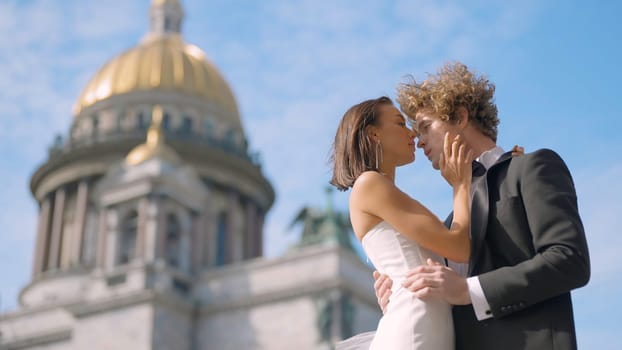 Wedding photo shoot. Action. A couple poses next to St. Isaac 's Cathedral , a young man with long curly hair and his bride with bare shoulders and long earrings.