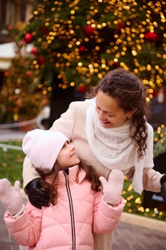 Authentic cheerful family, multi ethnic mother and little daughter smile, enjoying together happy winter holidays, standing by illuminated Christmas tree at traditional family market or Christmas fair