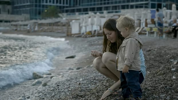 Mom and child throw stones into sea. Creative. Mother and child are relaxing at sea and playing on beach with stones. Mother and child are resting on shore throwing stones.