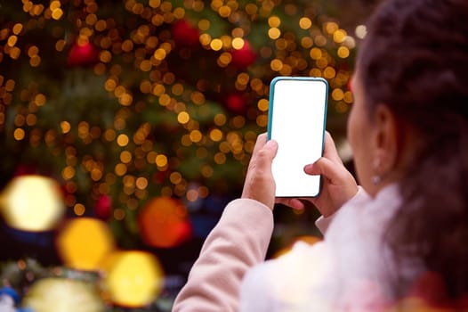 Close-up hands of a woman in winter clothes, using mobile phone, photographing the illuminated Christmas tree at Xmas fair, uploading mobile app, checking social media content on her modern smartphone