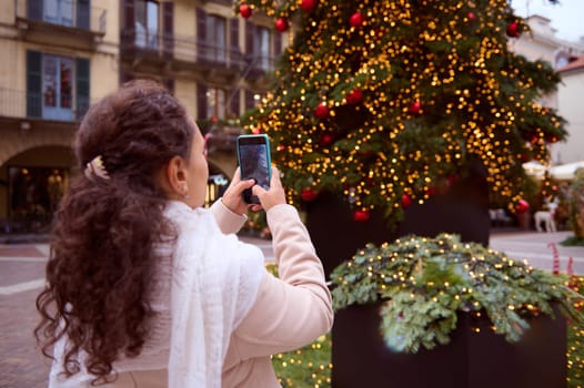 Rear view of a curly haired woman using mobile phone, photographing the illuminated Christmas tree while walking along medieval Italian city streets during Xmas holidays. Telephone in live view mode