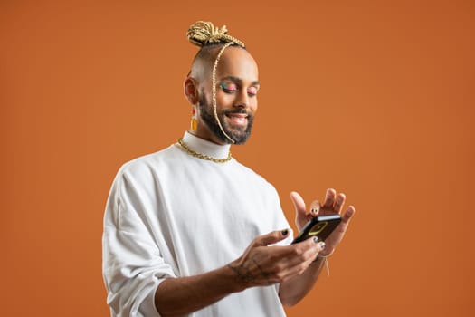 Latino gay man with bright makeup use smartphone isolated orange background. Confident Brazilian gay dressed white using mobile phone, sending message in social media or make online shopping