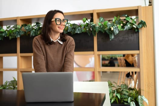 Woman with eyeglasses stands in an office at desk and looks away. Female dressed in smart casual stands near workplace. Businesswoman with eyeglasses working with laptop in creative office