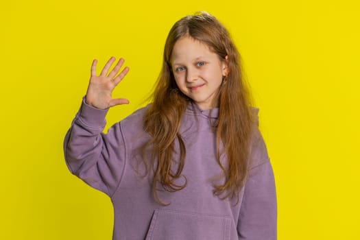 Young little redhead school girl smiling friendly at camera and waving hands gesturing hello or goodbye, welcoming with hospitable expression. Preteen female child kid isolated on yellow background