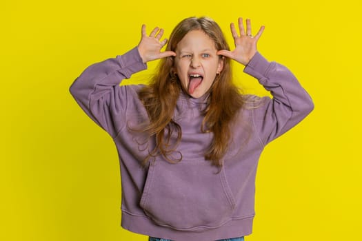 Cheerful funny bully young cute girl showing tongue making faces at camera, fooling around, joking, aping with silly face, teasing. Preteen redhead child kid on yellow background. Little children