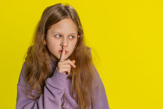 Shh be quiet please. Preteen child girl kid presses index finger to lips makes silence hush gesture sign do not tells gossip secret. Children isolated alone on studio yellow background. Copy-space
