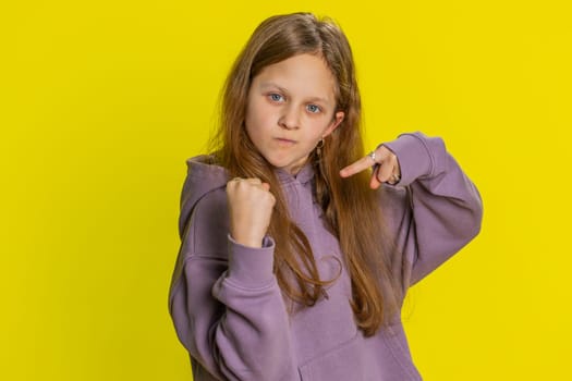 Portrait of aggressive angry young preteen child girl kid trying to fight at camera, shaking fist, boxing with expression, punishment, threaten, bullying abuse, mad fury. Children on yellow background