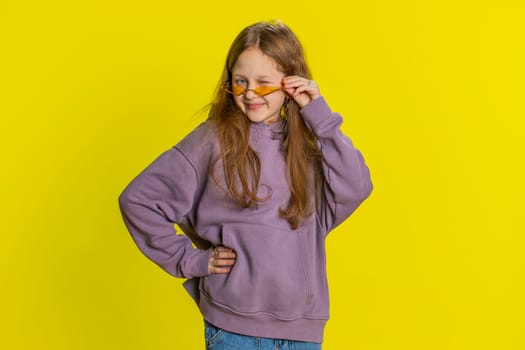 Playful happy young preteen child girl kid wearing orange sunglasses blinking eye, looking at camera with smile, winking, flirting, expressing optimism. Teenager little children on yellow background