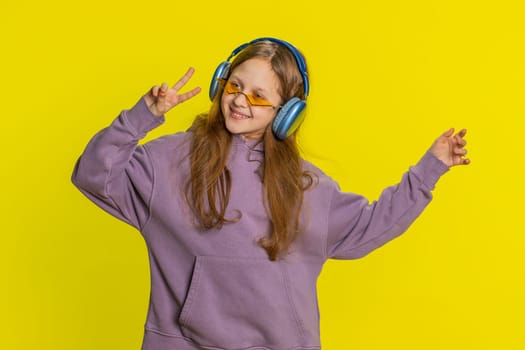 Happy smiling preteen child girl in headphones listening music, dancing disco, fooling around having fun expressive gesticulating hands relaxing on party, making funny moves. Kid on yellow background