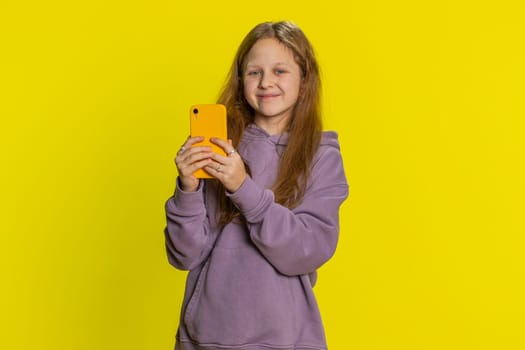 Smiling preteen child girl kid using mobile smartphone typing new post on web, looking at camera, sms message, watching video addiction of social media networks. Teenager children on yellow background