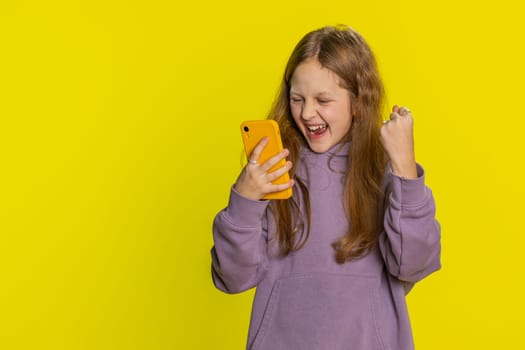 Young preteen child girl kid use mobile smartphone typing browsing say Wow yes found out great big win good news celebrate doing winner gesture. Little cute children on studio yellow background