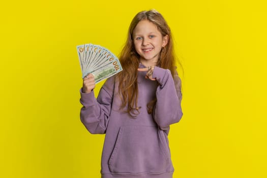 Portrait of rich pleased young cute school girl waving money dollar cash banknotes bills like a fan success lottery winner big income, wealth. Teen redhead female child kid on studio yellow background