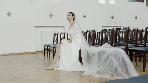 Brunette bride in fashion white wedding dress with makeup. Action. Wedding day of bride, woman sitting on a chair in front of empty rows