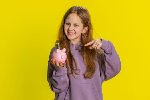 Young preteen child girl kid putting dollar pocket money cash into piggy bank saving for future education. Teenager children with coins box finance income growth, economy on yellow background indoors
