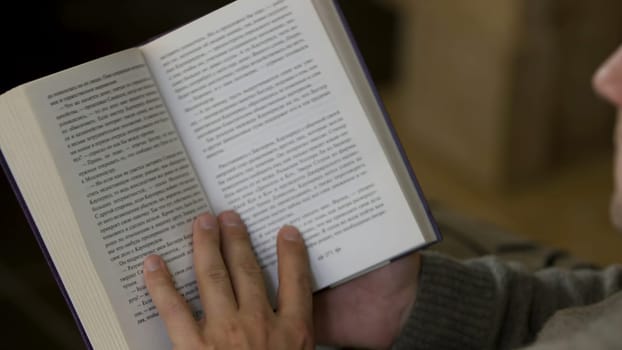 Over the shoulder view of a man reading a book at home. Middle aged male sitting and reading a paper book, turning pages, relaxing at home.