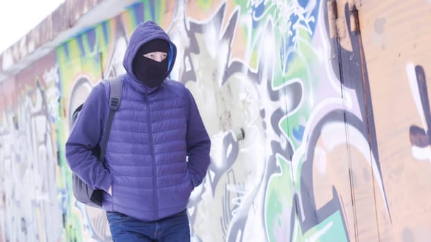 Serious young man in casual outfit and balaclava walking along the grafitti wall. Male with a backpack hiding his face and looking around, concept of vandalism.