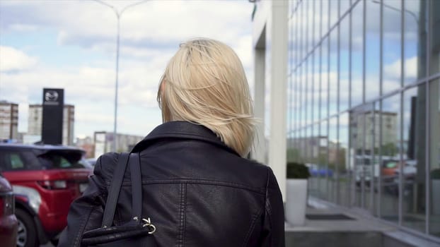 Rear view of a blonde in a black leather jacket walking in the city street. Young, blonde woman walking through the streets of the city with a bag over her shoulder.