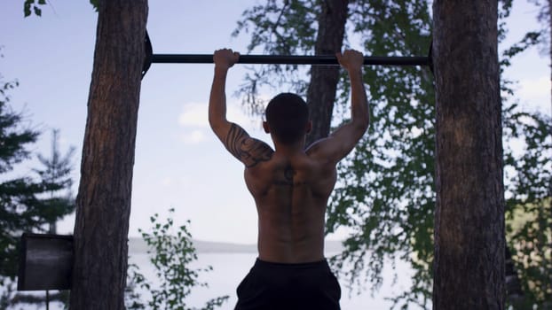 Back view of tattooed man doing pull-ups while having workout in picturesque autumn park. Young athlete in the tattoo pulled on the bar in the Park.
